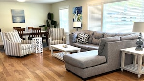 A living room with hardwood flooring, a gray sofa, striped armchairs, a coffee table, and a dining area with a table and chairs.