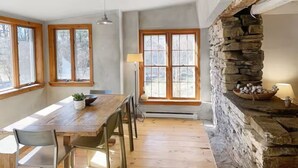 A dining area with a wooden table and chairs, large windows, and a stone fireplace.