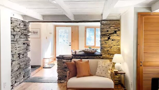 A cozy interior with a stone wall, wooden floors, a sofa, and a kitchen area with a stone wall and windows.