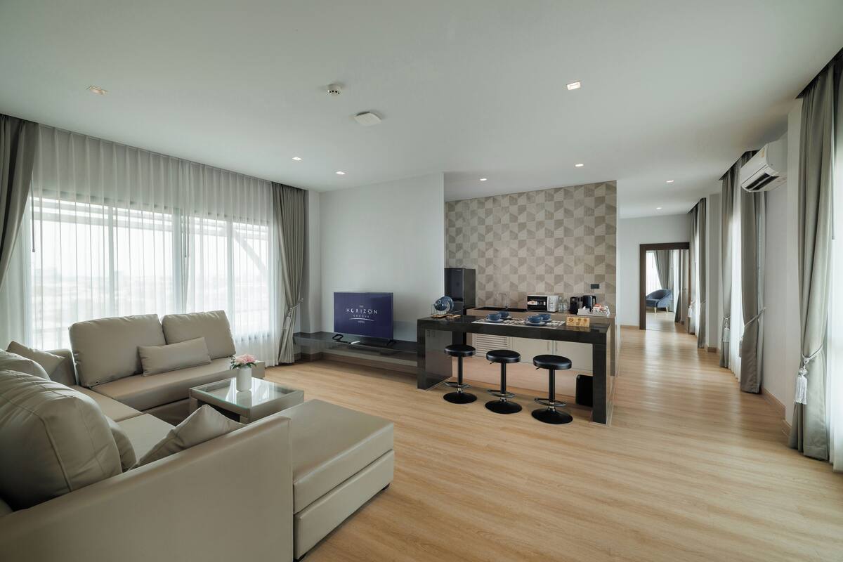 Een moderne woonkamer met een bank, salontafel en televisie. Er is een keukenhoek met een bar en krukken. De kamer heeft houten vloeren en grote ramen met gordijnen.