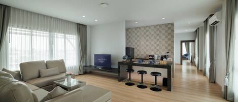 Una sala de estar moderna con un sofá, una mesa de centro y un televisor. Hay una zona de cocina con una barra y taburetes. La habitación tiene suelo de madera y ventanas grandes con cortinas.