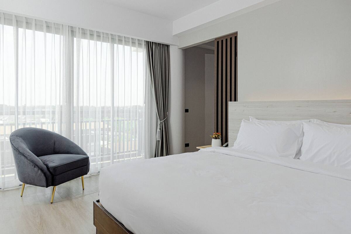 Een moderne hotelkamer met een groot bed, een grijze fauteuil en uitzicht op de stad door dunne gordijnen.