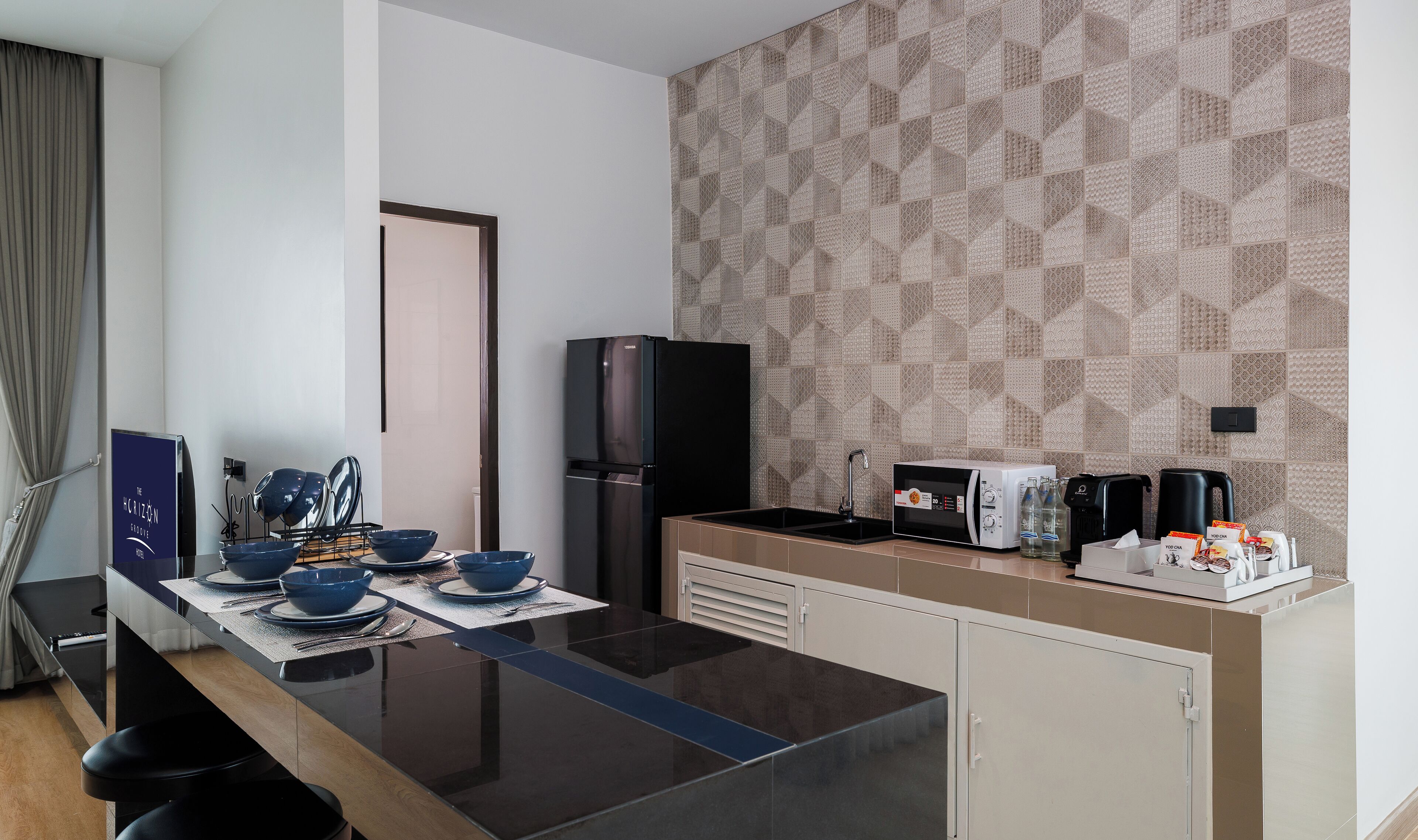 A modern kitchen with a black refrigerator, a microwave, and a coffee maker. There are blue bowls and plates on the counter, and a sign for The Horizon Groove Hotel.