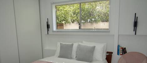 A bedroom with a bed, a chair, a window with blinds, and a wall-mounted shelf with books.
