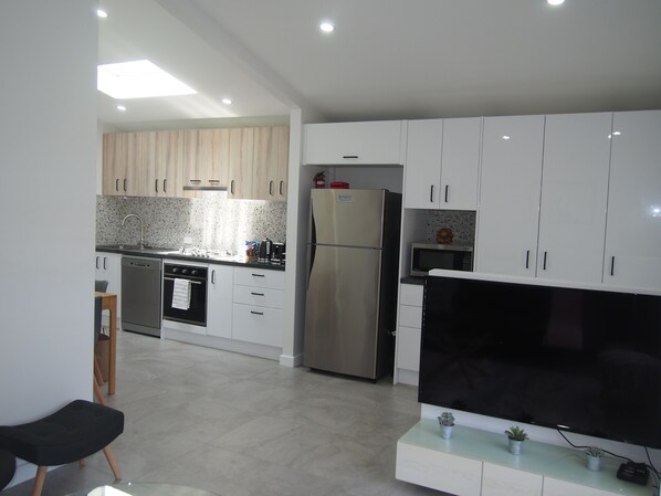 A modern kitchen with white cabinets, a stainless steel refrigerator, a microwave, and a large flat-screen TV on a glass table.