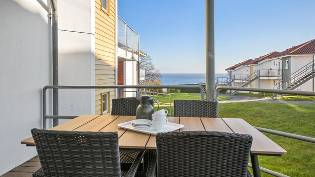 A balcony with a table set for two, overlooking a grassy area and a building with a red roof. - Gudhjem Søpark Lejl. 29 (Gudhjem)