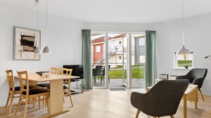 A modern living room with a dining area, large windows, and a view of a residential building. - Gudhjem Søpark Lejl. 29 (Gudhjem)