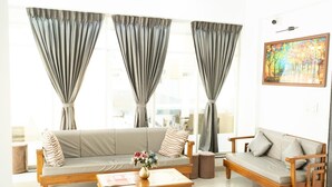 A modern living room with a sofa, a wooden coffee table, and a side table. There are two large windows with curtains, and a painting on the wall. - Sunny Executive Satara (Satara)