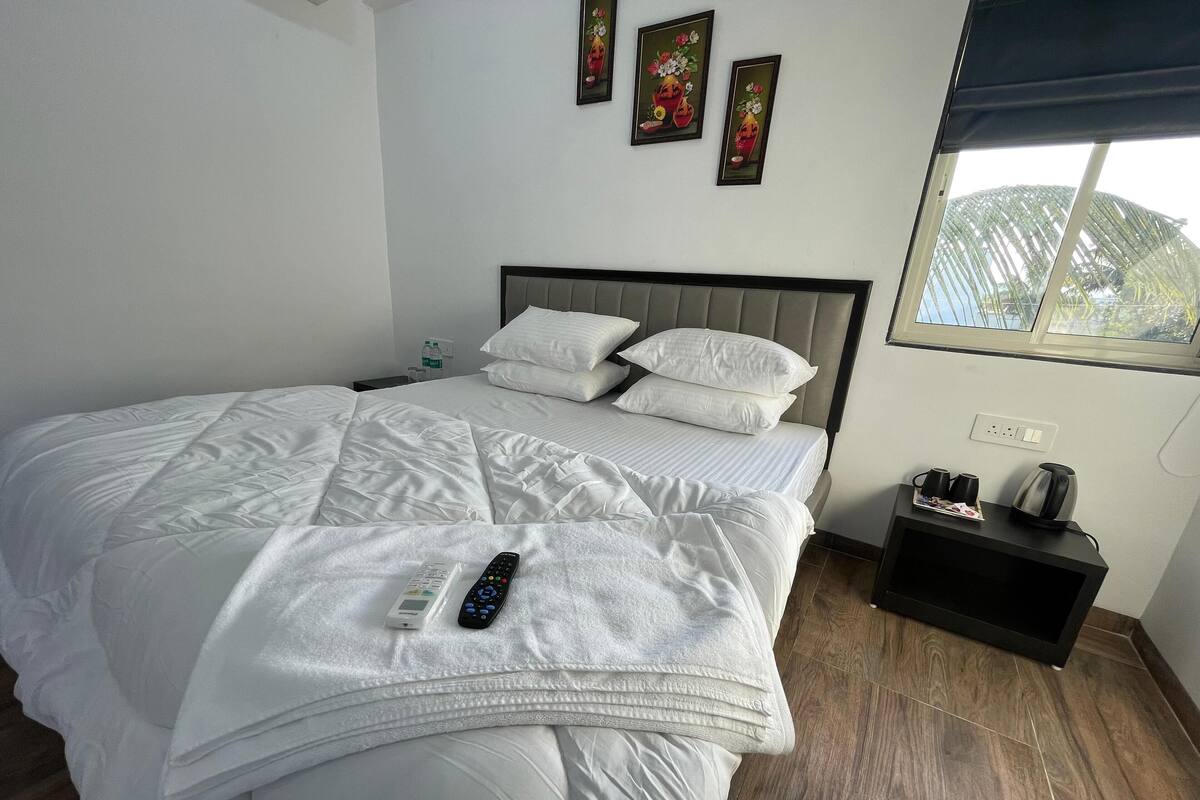 A neatly made bed with white linens, a remote control, and a bedside table with a kettle and cups.