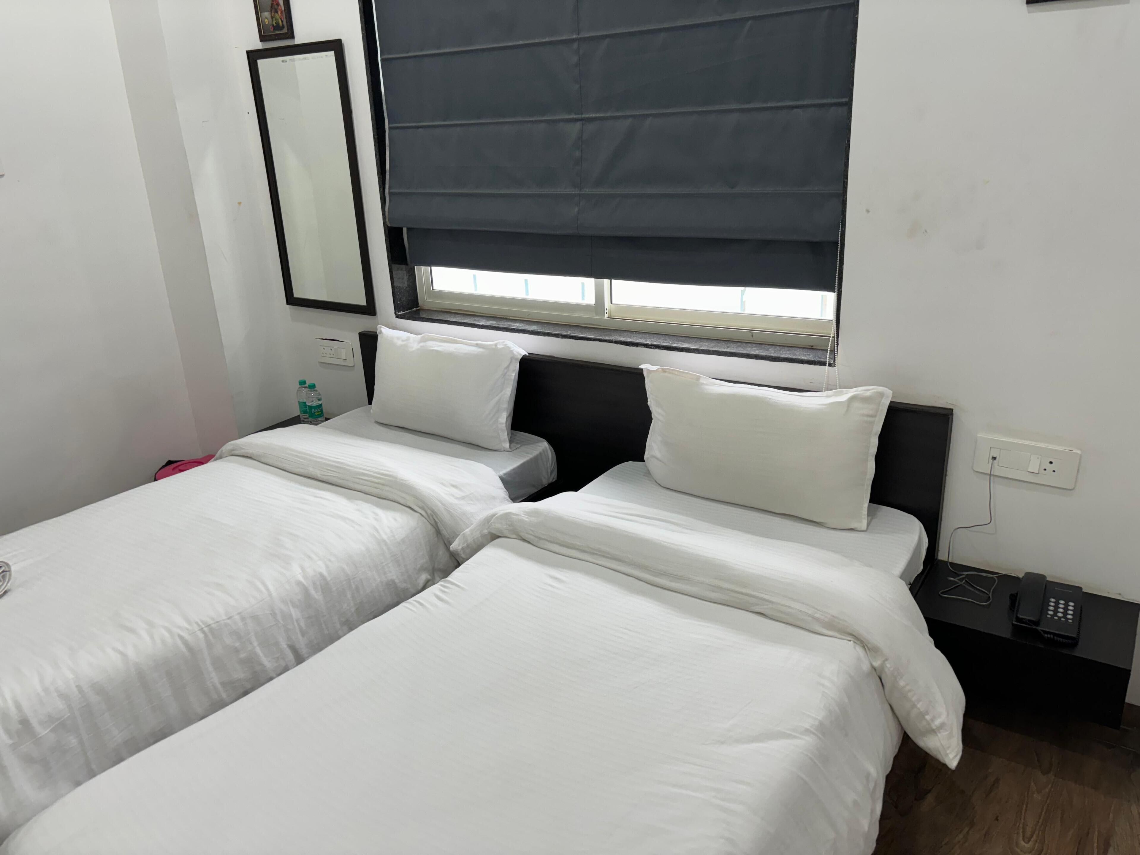 A hotel room with two single beds, a window with a dark blind, a bedside table with a phone and charger, and a wall-mounted mirror.