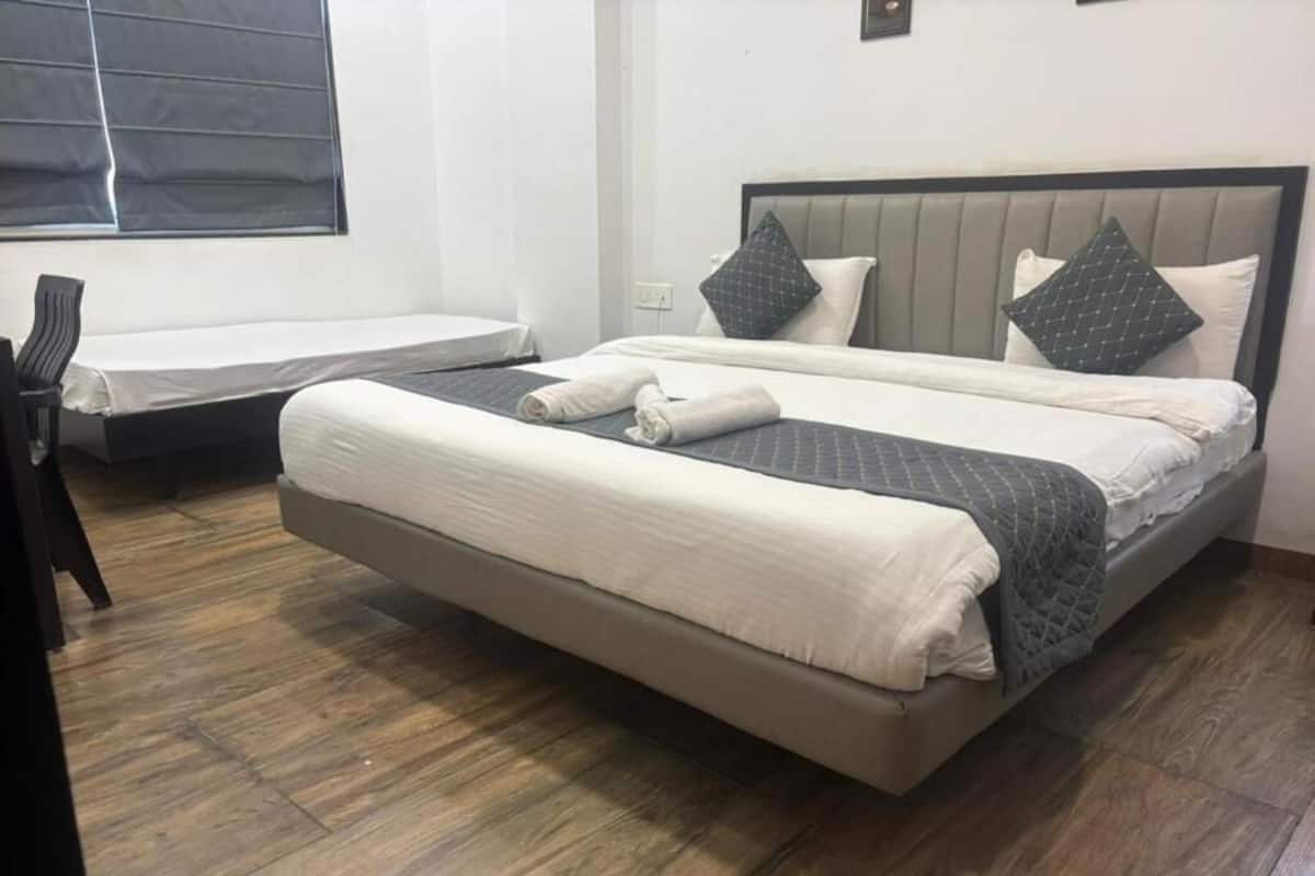 A double bed with a grey headboard and white bedding, a small bedside table, and a window with blinds.