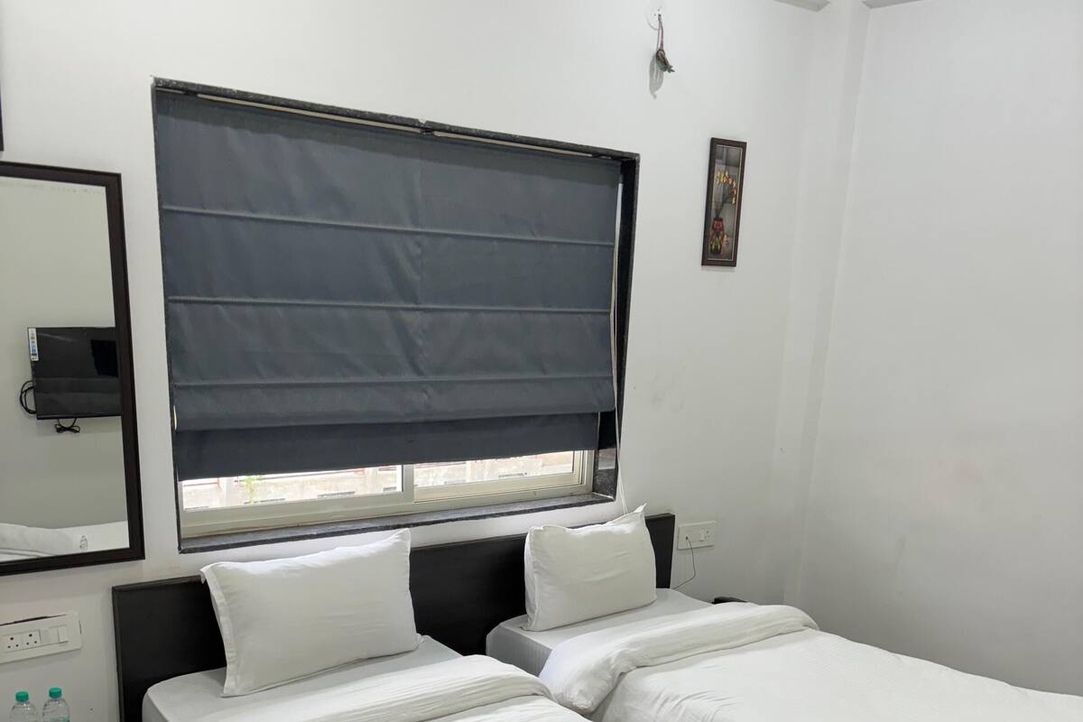 A hotel room with two single beds, a window with a dark blind, a small bedside table with a mirror and a TV, and a wooden floor.