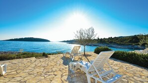 A stone patio with two white lounge chairs facing a body of water and a tree. - Nice apartment in Rogoznica with sauna (Rogoznica)