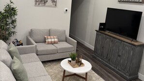 Un salon avec un canapé gris, une table basse, un téléviseur posé sur un meuble en bois, un ventilateur de plafond et un panneau décoratif représentant le mot « FAMILLE » accroché au mur.
