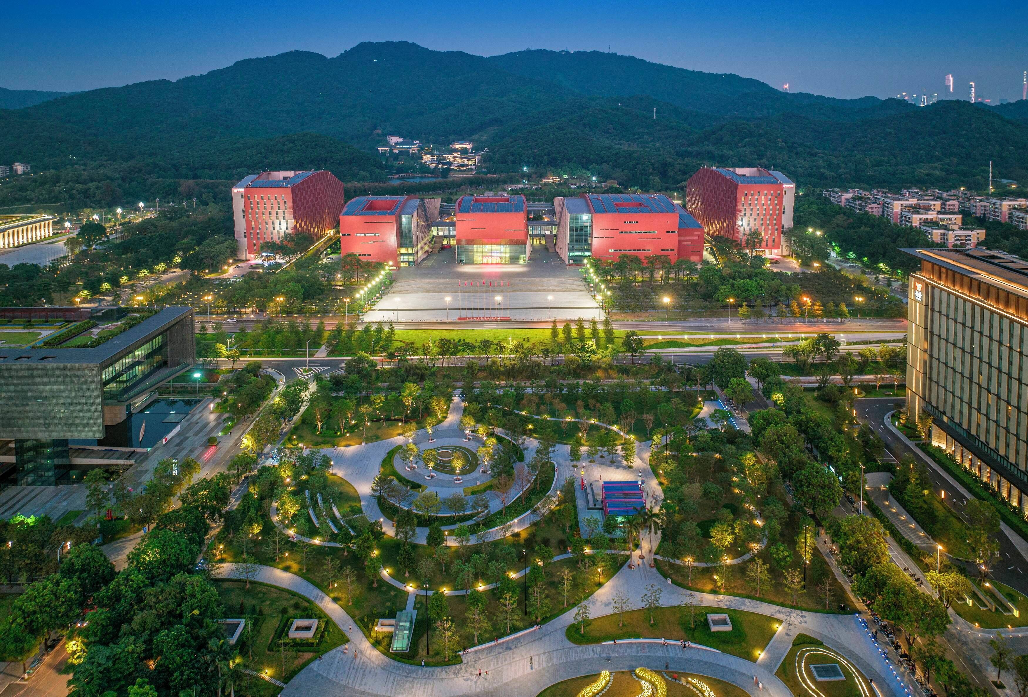 A modern urban area with a central plaza, surrounded by buildings with red roofs, and a mountainous backdrop.