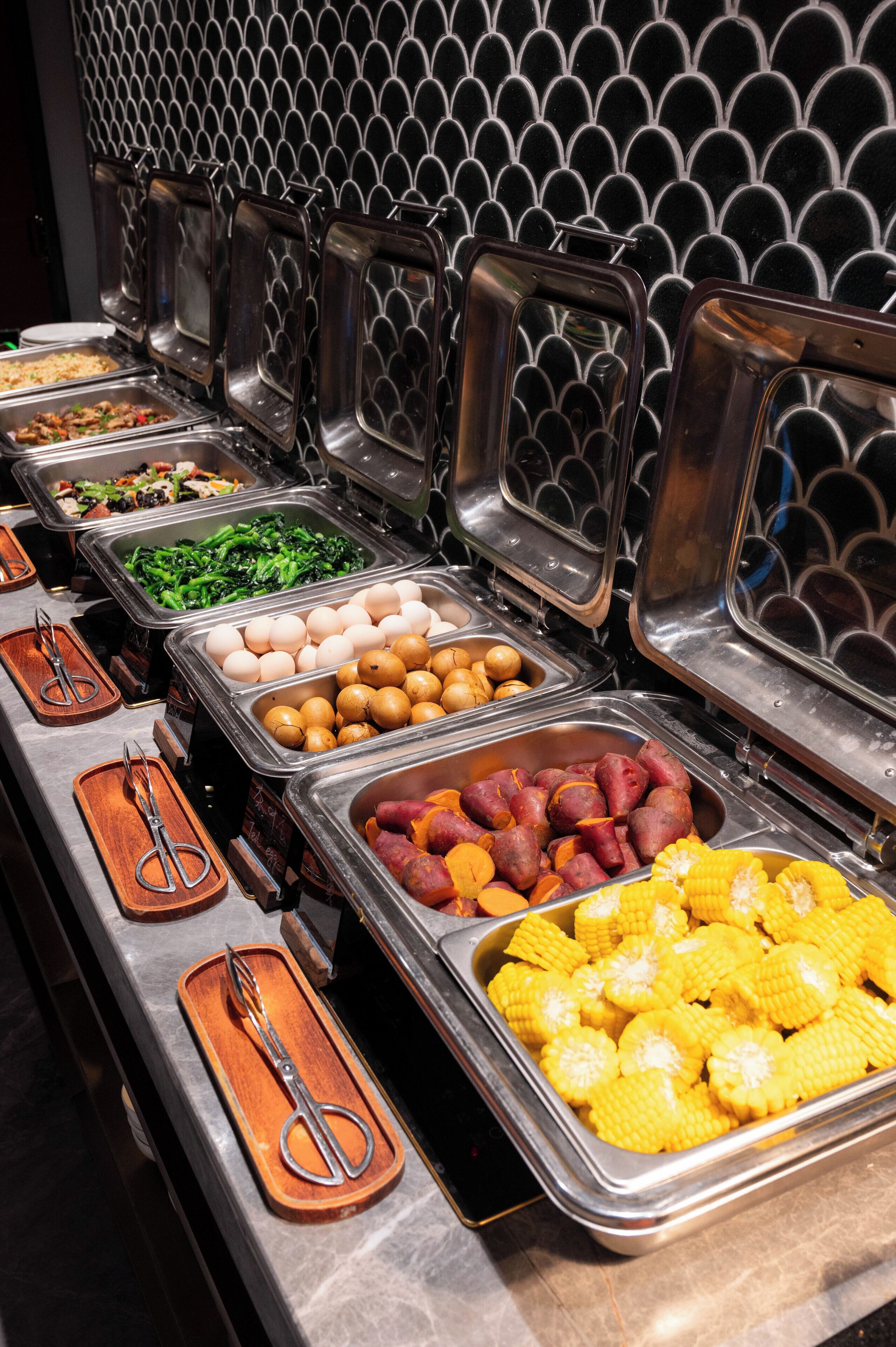 A buffet with various food items in metal containers, including vegetables, eggs, and corn.