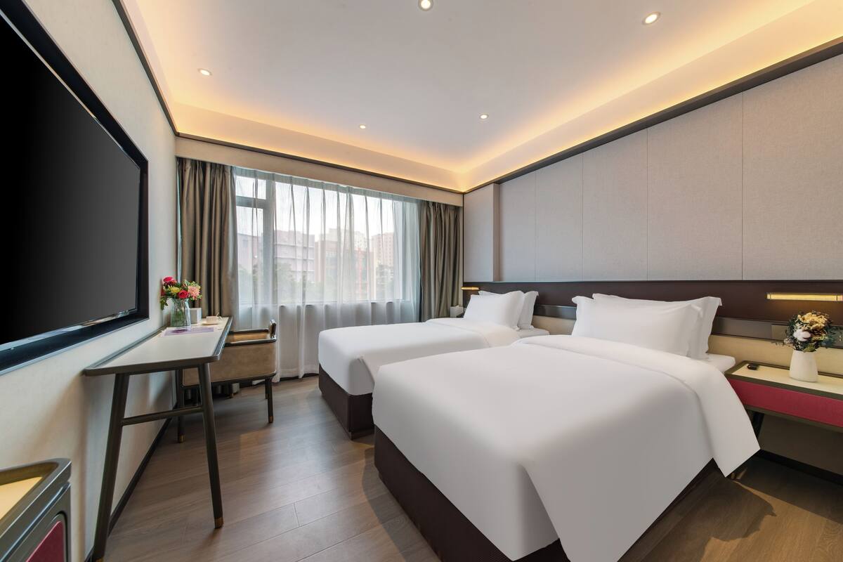 A hotel room with two beds, a desk, a TV, and a window with curtains.