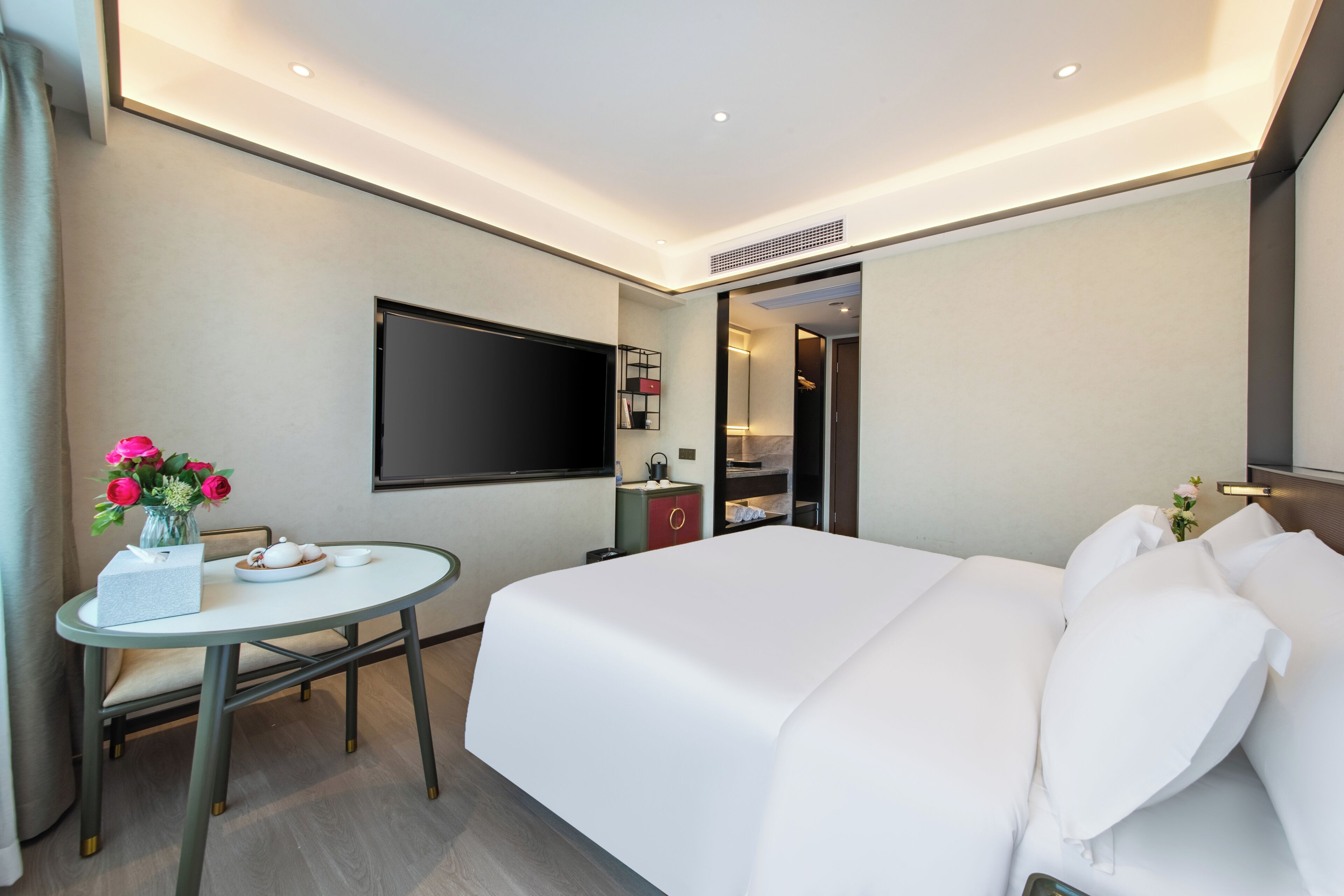 A hotel room with a bed, a TV, a small table with a vase of flowers, and a desk with a chair.