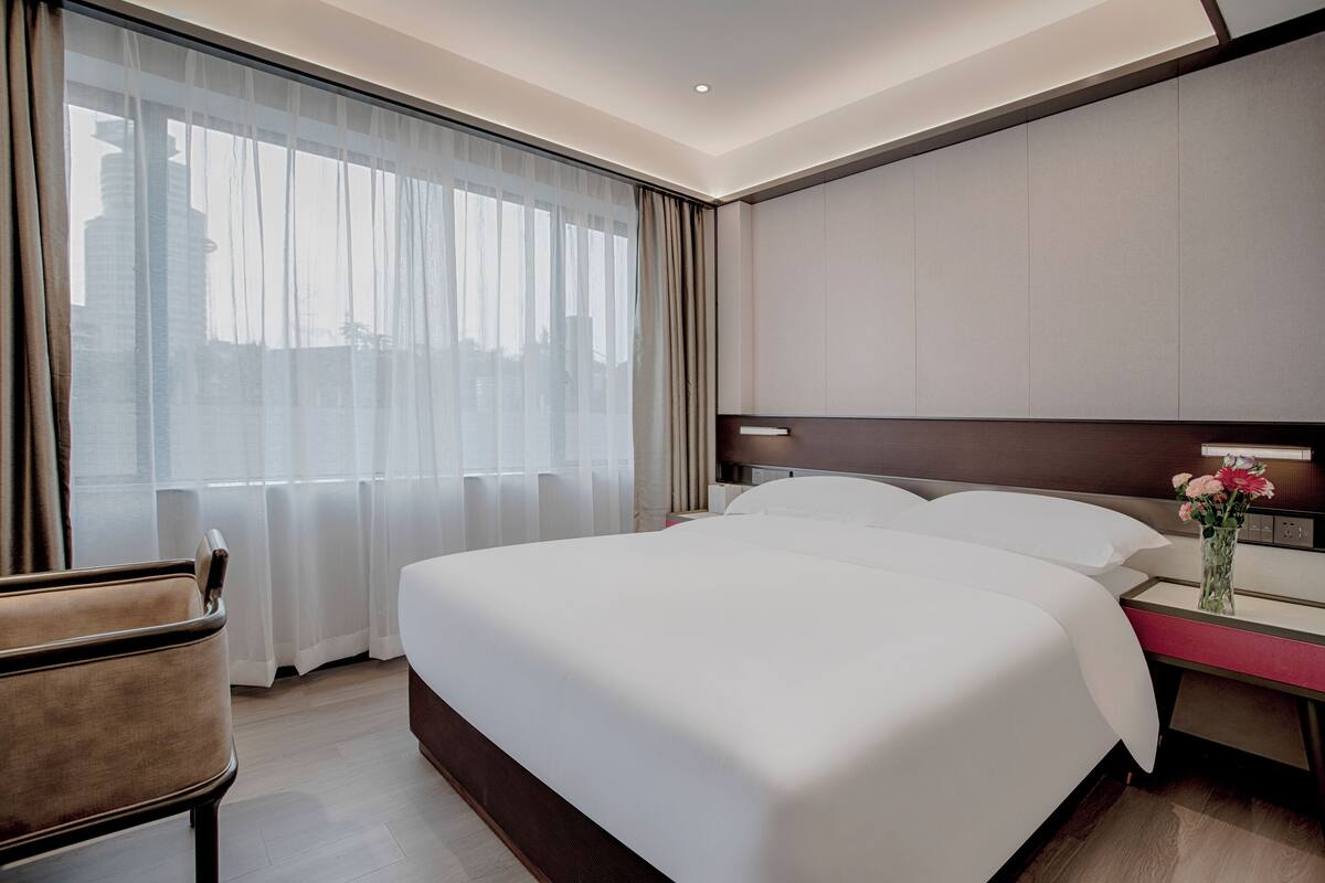 A hotel room with a large bed, a bedside table with a vase of flowers, a chair, and a large window with curtains.