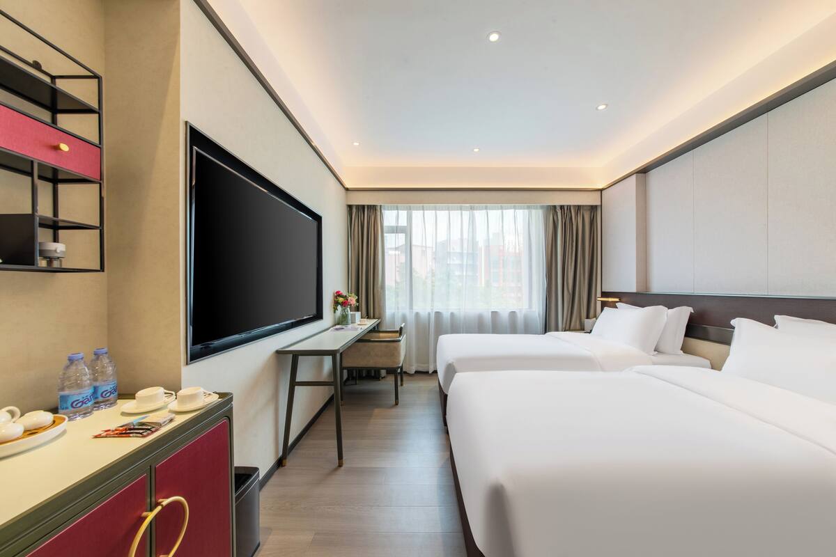 A hotel room with two beds, a desk with a flat-screen TV, a minibar, and a large window with curtains.