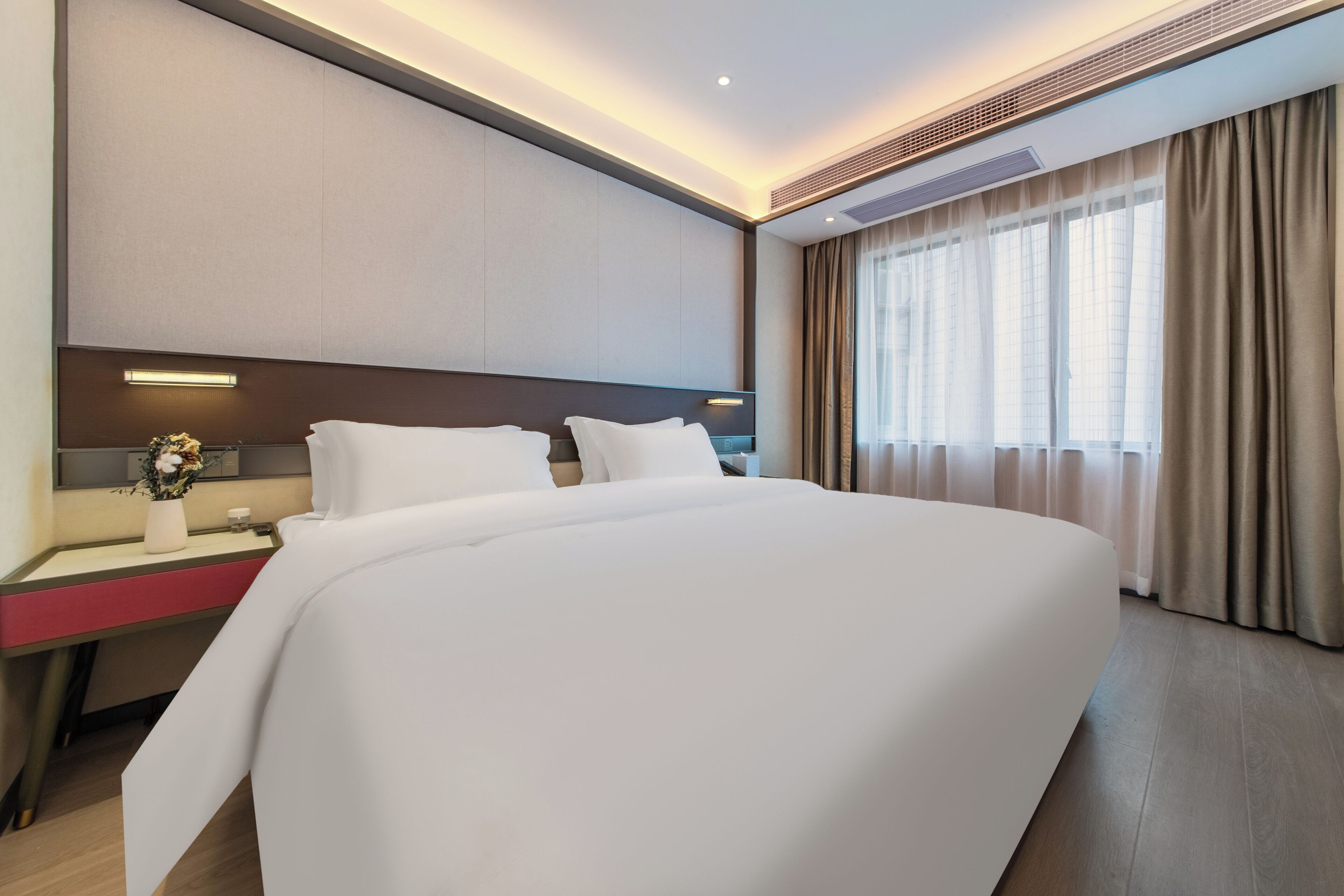 A modern hotel room with a large bed, a bedside table with a vase of flowers, a desk, and a large window with curtains.