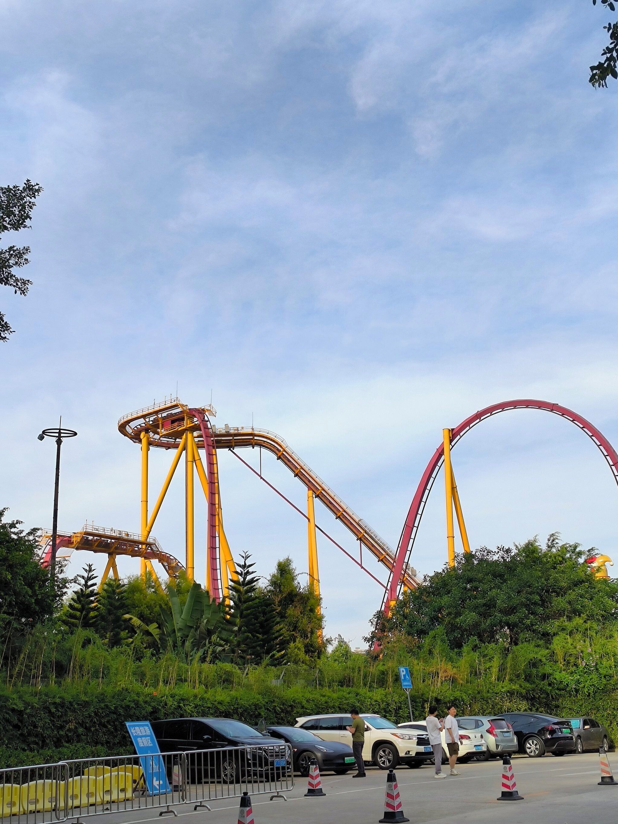 A roller coaster with a yellow and red track, surrounded by greenery and parked cars.