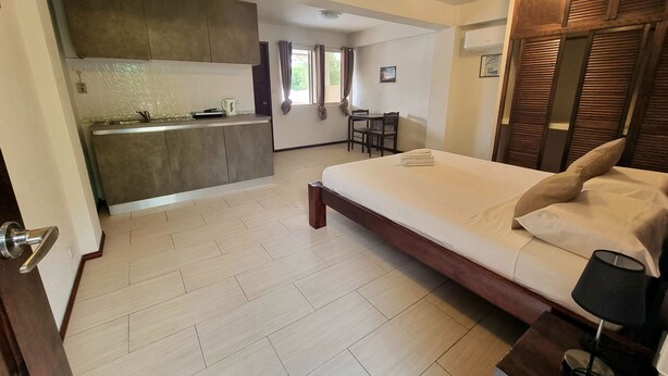 A bedroom with a large bed, a bedside table, and a nightstand. There is a small kitchenette in the background with a sink and a stove. - Day & Night Accommodations (Daanbantayan)
