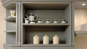 Eine Küche mit einem grauen Schrank, der weiße Teetassen sowie Zucker- und Kaffeebohnenbehälter enthält. Eine schwarze Wasserkochkessel steht auf der darunterliegenden Arbeitsfläche.