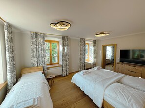 A hotel room with two beds, a large window with curtains, a radiator, a TV on a wooden cabinet, and a ceiling light fixture.