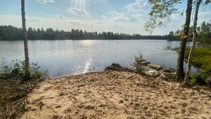 Une plage de sable bordée d’un lac paisible, entourée d’arbres et sous un ciel dégagé.
