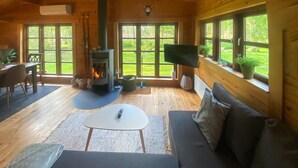 L’intérieur chaleureux d’un chalet avec un poêle à bois, un canapé, une table à manger et de grandes fenêtres donnant sur le paysage extérieur.