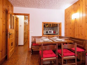 A dining area with wooden walls, a kitchen visible through a window, and a door leading to another room.