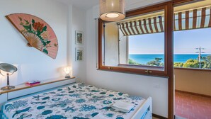Ein Schlafzimmer mit Bett, Nachttischlampe, dekorativem Fächer und einem Fenster mit Meerblick.