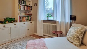 Une chambre à coucher comprenant un lit, une table de chevet, un radiateur, une bibliothèque et une fenêtre avec des rideaux.