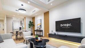 A modern living room with a flat-screen TV, a dining area, and a kitchen in the background.