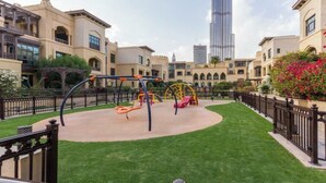 A playground with swings and a climbing structure, surrounded by well-maintained grass and a fence, with a tall building in the background.