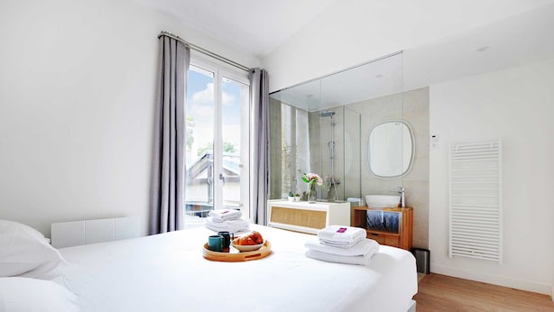 A hotel room with a large bed, a bedside table with a tray of breakfast items, a bathroom with a mirror and sink, and a view of the outside. - Beautiful Apartment- 3br/10p - Montparnasse (Paris)