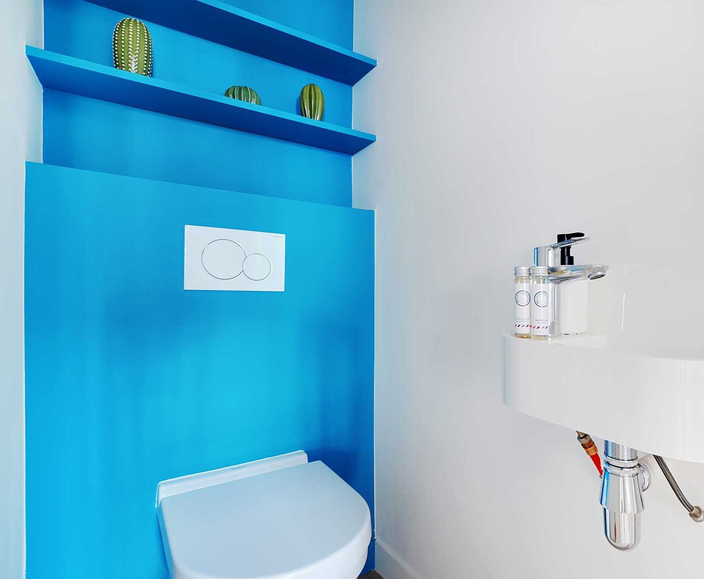A bathroom with a blue accent wall, a white toilet, a shelf with decorative items, and a wall-mounted faucet with a soap dispenser.