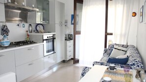 A modern kitchen with white cabinets, a stove, and an oven. There is a dining area with a table and chairs, and a sofa with a patterned cover. - Apartment with rooftop swimming pool (Bibione)