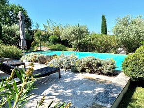 A pool area with lounge chairs and a covered umbrella, surrounded by lush greenery and a clear blue sky. - Villa infinity pool near Lourmarin (Lauris)