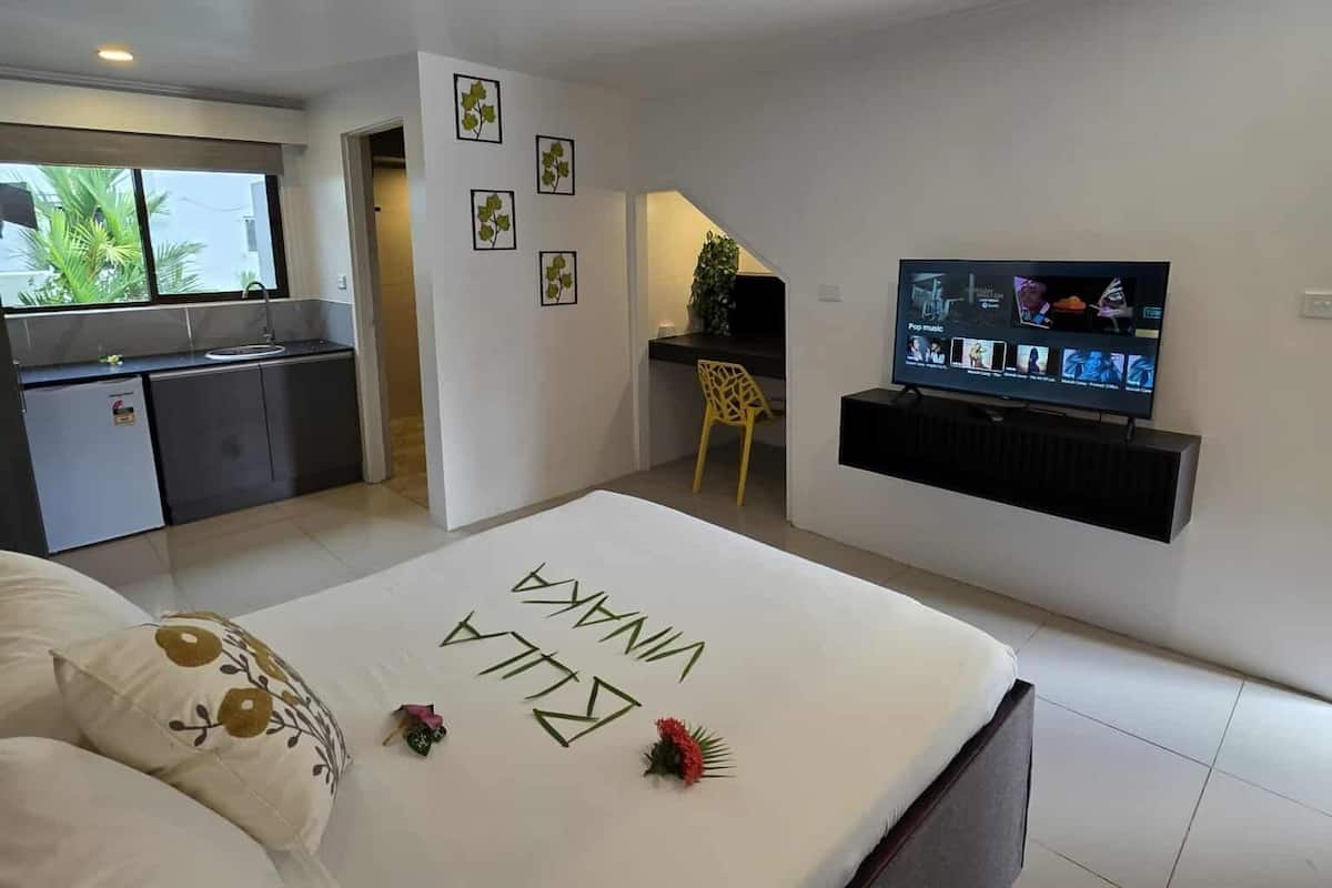 A modern bedroom with a bed, a TV, and a kitchen area visible through an open door.