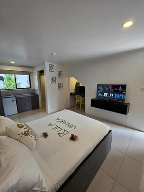 A modern bedroom with a bed, a TV, and a kitchen area visible through an open door.