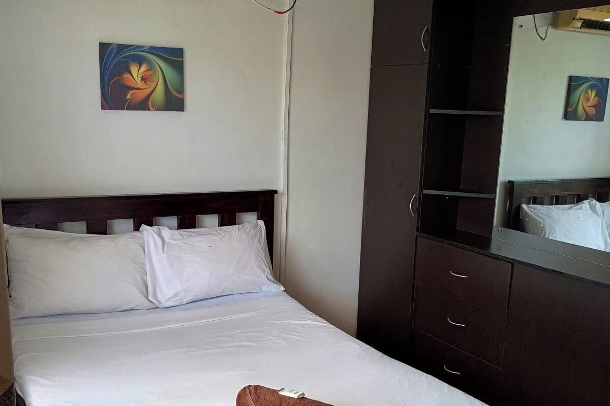 A neatly made bed with white linens and a brown towel, a dark wooden wardrobe, and a wall-mounted air conditioner.