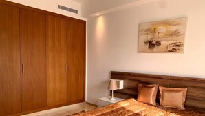 A bedroom with a large wooden wardrobe, a bed with a patterned bedspread, a nightstand with a lamp, and a painting on the wall.