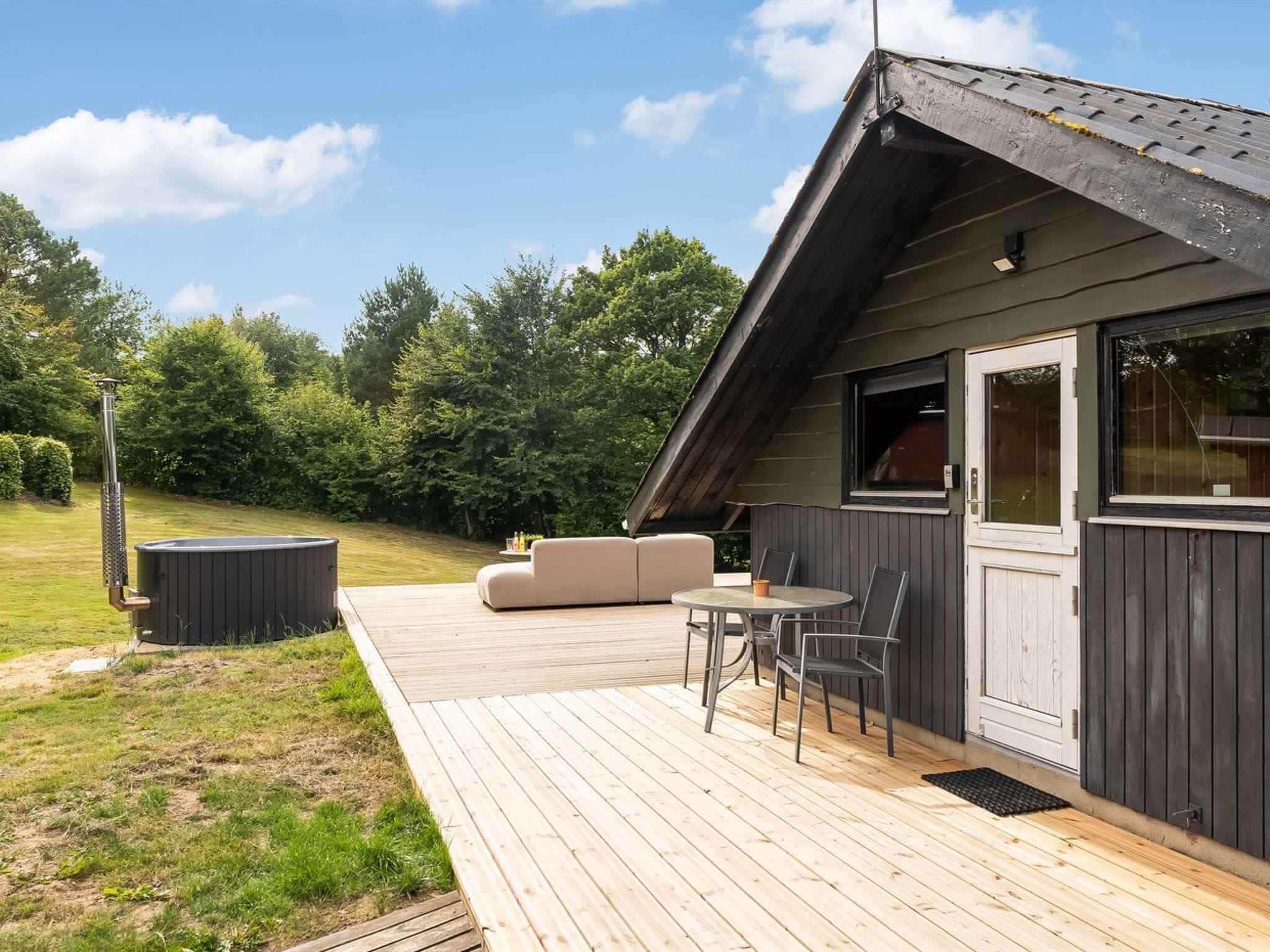 Eine Holzterrasse mit einer Sitzecke, Tisch, Whirlpool und überdachtem Außenbereich mit Blick auf Bäume.