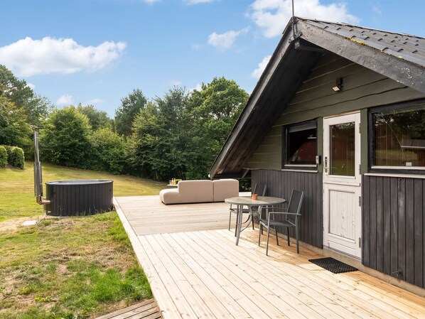 A wooden deck with a sofa and table, a hot tub, and a covered outdoor area with a view of trees.