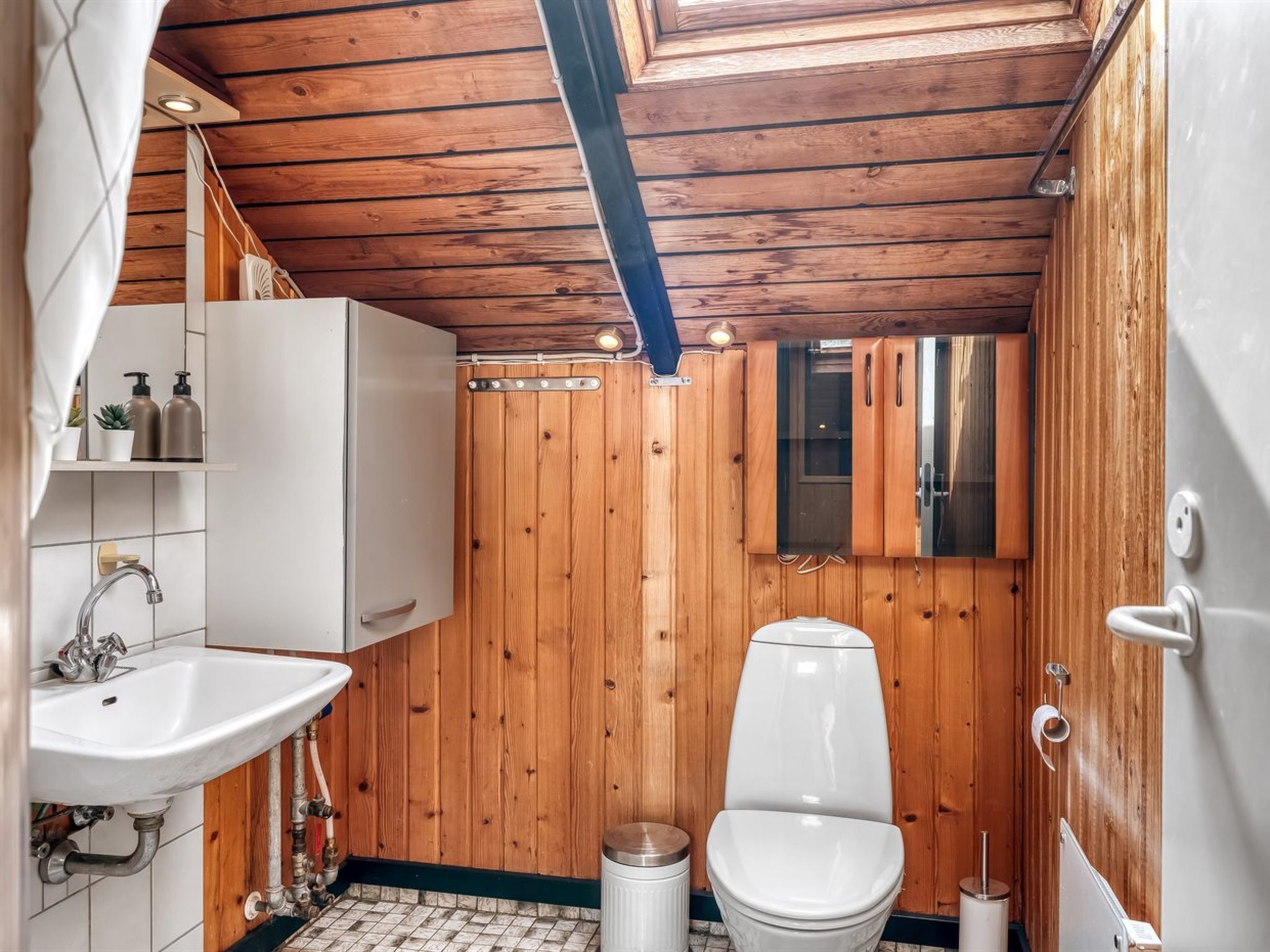 Et lille badeværelse med træpaneler, en hvid vask, toilet og spejl.