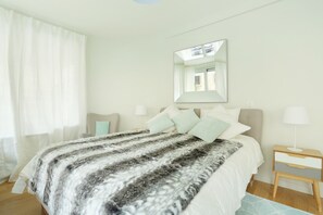 Un dormitorio con cama, mesita de noche, espejo y ventana con cortinas.