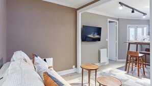 Een moderne woonkamer met een bank, twee houten bijzettafels, een flatscreen televisie aan de muur en een eethoek met een tafel en stoelen.