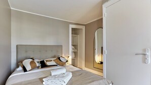 Een net opgemaakt bed met hoofdbord, een nachtkastje met lamp en een garderobe met spiegel.
